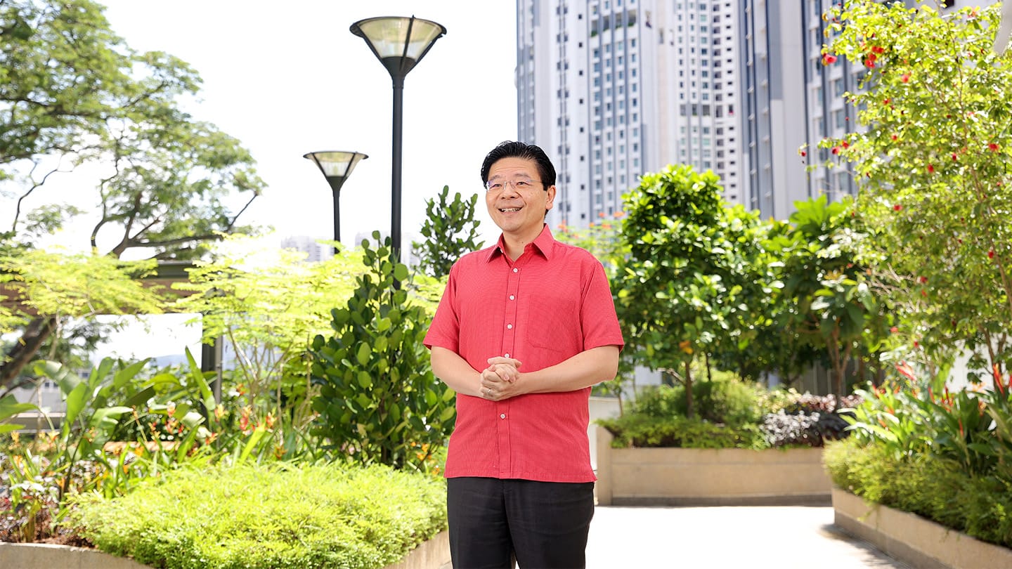 Asian man in a red shirt stands in a garden with city buildings and lampposts.